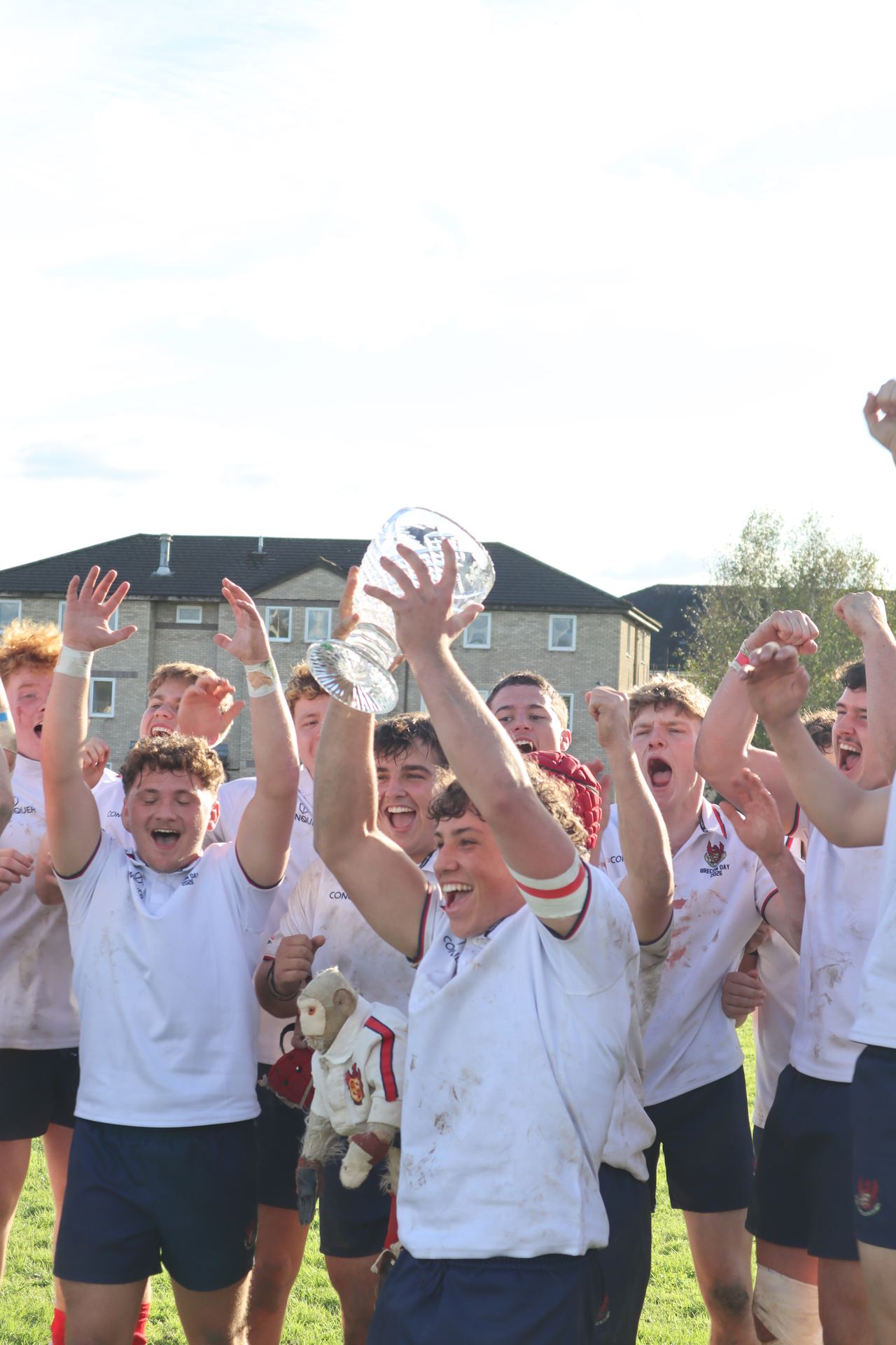 The 1st XV celebrating on the pitch at Tredegar Close, the captain raising the trophy with the College Monkey beside him