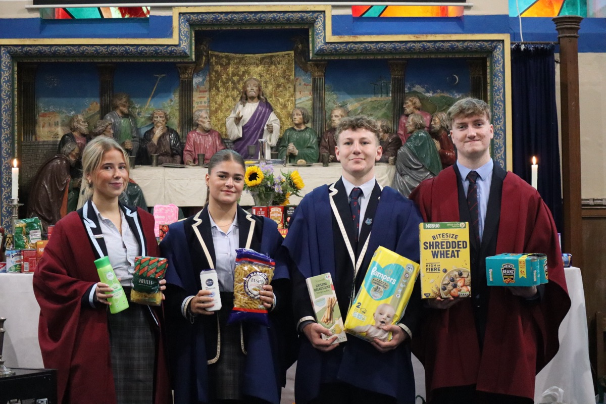 Four Senior School Prefects in gowns holding food and household items from the Harvest collection at the Chapel altar